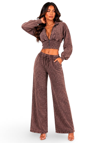 A woman modeling a matching two-piece lounge set in a washed brown color, featuring a cropped zip-up hoodie with long sleeves and matching wide-leg drawstring pants, set against a plain white background.