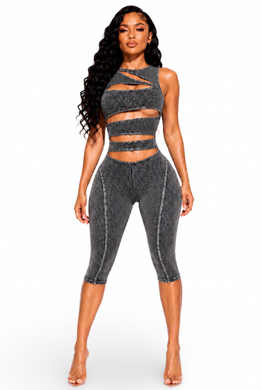 A woman modeling a matching two-piece activewear set in a washed charcoal gray color. The set includes a strappy cutout crop top and high-waisted cropped leggings with vertical seam detailing, shown against a plain white background.