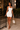 A woman models a fitted white halter-neck mini dress in an outdoor patio setting at night, holding a matching white handbag.