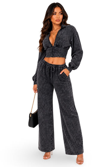 A woman modeling a dark gray acid-wash two-piece set consisting of a cropped zip-up hoodie and matching wide-leg drawstring pants, paired with clear high-heeled sandals and a small black handbag, against a plain white background.