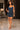 A woman modeling a fitted mini dress made from blue denim and dark gray fabric panels, photographed in a modern indoor showroom with a marble floor and dark curtains in the background.