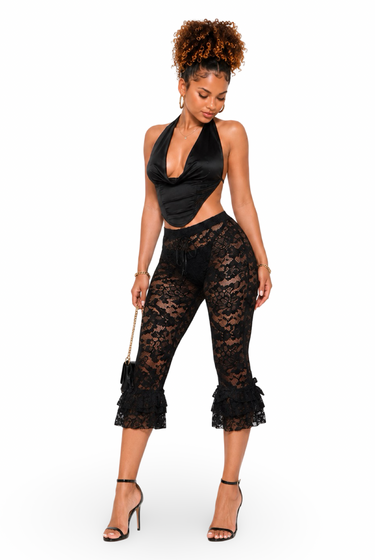 A woman modeling a black lace two-piece outfit consisting of a halter crop top and matching high-waisted flared cropped pants with ruffled hems, paired with black high-heeled sandals against a plain white background.
