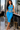 A woman models a bright blue two-piece set consisting of a sleeveless cutout crop top and matching high-waisted leggings, standing in a modern interior with a city skyline view in the background.