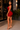 A woman posing in a tight, sleeveless red mini dress with clear high-heeled sandals in an outdoor patio setting at night.