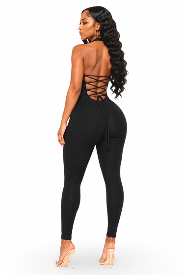 A woman modeling a black one-piece jumpsuit from the back, featuring a lace-up back design, on a solid white background.